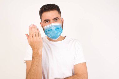 What the hell are you talking about, nonsense. Studio shot of frustrated male with blond hair gesturing with raised palm, frowning, being displeased and confused with dumb question over gray wall.  Caucasian man wearing medical mask 