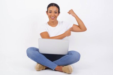 Waist up shot of blonde woman raises hand to show her muscles, feels confident in victory, looks strong and independent, smiles positively at camera, stands against gray background. Sport concept. Young beautiful Arab woman using laptop computer clipart