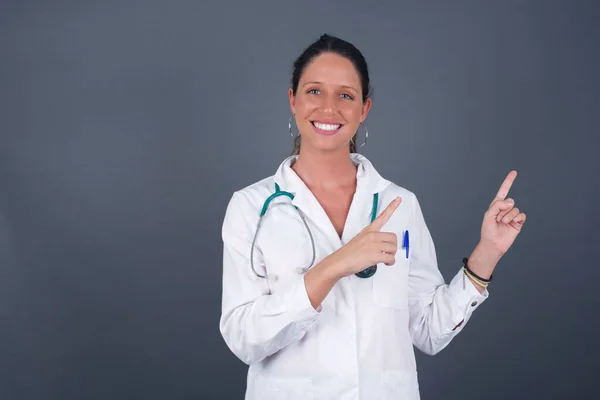 广告概念 一个正面的 漂亮的 高加索裔的医生女人 在灰蒙蒙的背景下 面带微笑 与灰蒙蒙的背景隔离 用她的两个食指指尖着 望着空白的复制空间 — 图库照片