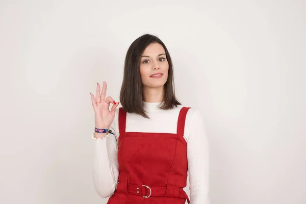 Glad attractive woman shows ok sign with hand as expresses approval, has cheerful expression, being optimistic. Standing against white wall.