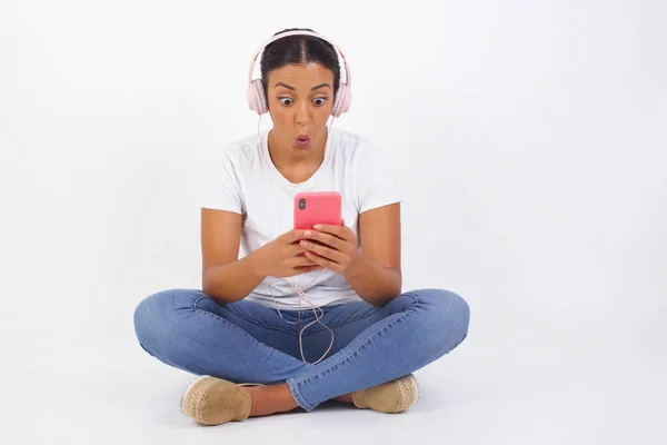 Horizontal shot of pretty young female isolated over pink background, looks with bugged eyes, holds modern smart phone, receives unexpected message from friend, reads reminder.