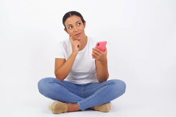 Image of a thinking dreaming young beautiful woman posing isolated over pink wall background using mobile phone.