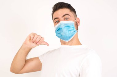 Neşeli, yakışıklı, Avrupalı koyu renk saçlı, mavi gözlü adam neşeli, memnun ve kendinden emin görünüyor. Başparmağıyla işaret ediyor. Gri stüdyo arka planında izole edilmiş. Tıbbi maske takan beyaz bir adam.