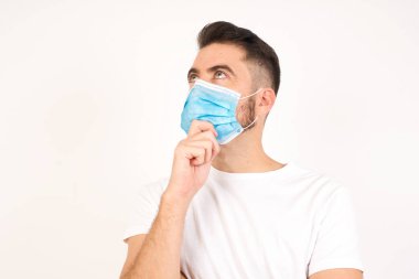 Tıbbi maske takan düşünceli gülümseyen bir adamın portresi elini çenesinin altında tutar, yan bakar, ilginç bir şey düşünür ya da merak eder, sıradan giyinir, gri stüdyoya karşı poz verir. Karar alma kavramı.