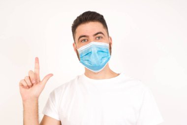 Hoş görünümlü mavi gözlü genç adam modelinin yakın portresi zekice bir ifadeye sahiptir, bir parmağını kaldırır, önemli bir şeyi söylemeyi unutmaması gerektiğini hatırlar. Tıbbi maske takan beyaz bir adam. 