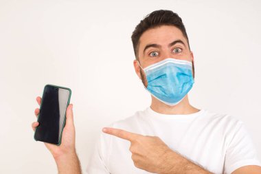 Tıbbi maske takmış, beyaz arka planda dikilip dijital cihazı işaret eden heyecanlı bir beyaz adam. Stüdyoda, elinde boş ekranla akıllı telefon tutan adamın şok olmuş görüntüsü. Reklam kavramı.