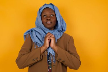 Tatlı yüzlü, rüya gibi, Afro-Amerikan Müslüman bir kadın. Elleri yüzü kapalı, hoş bir şey için heyecanlı, gri arka planda poz veriyor..