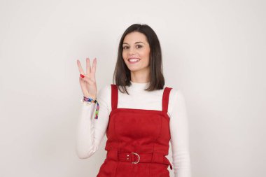 Gri duvara yaslanmış genç bir kadın üç numarayı gösterip işaret ederken kendinden emin ve mutlu bir şekilde gülümsüyor..