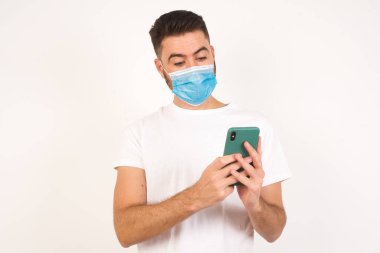 Tıbbi maske takan yakışıklı beyaz bir adamın yatay görüntüsü, beyaz arka plan üzerinden izole edilmiş, böcek gözlü, modern akıllı telefonu elinde tutan, arkadaşından beklenmedik bir mesaj alan,.
