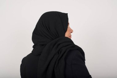 Siyah tesettürlü, uzun düz dalgalı ve parlak saçlı, gri duvarda duran Müslüman bir kadının arka tarafı. Stüdyo Çekimi. 