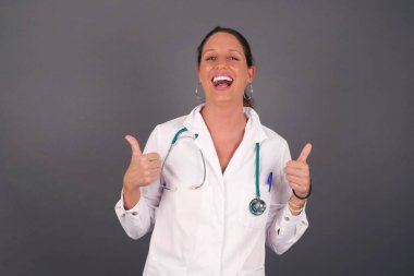 Gri arka planda izole edilmiş, olumlu içerik ve mutluluk duyguları gösteren genç iyimser kadın doktorların yakın çekimi. Kalite ve tavsiye ile tatmin kavramı.