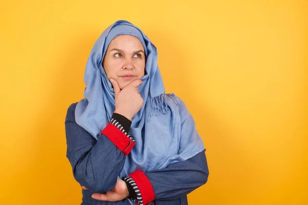 ヒジャーブのスタイリッシュな中年イスラム教徒女性の肖像画は 疑わしく懐疑的な表現 心配と疑念を持って手と横を見ています 背景の上に屋内で立っている — ストック写真