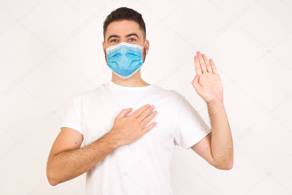 I swear, promise you not regret. Portrait of happy, sincere young man wearing medical mask raising one arm and hold hand on heart as give oath, pledge, telling truth, want you to believe, standing over yellow background