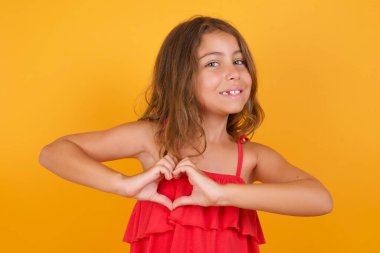 Sarı arka planda kalbi gösteren kırmızı elbiseli sevimli küçük bir kız. 
