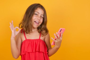 Stüdyonun sarı arka planında kırmızı elbise giyen küçük güzel bir kız. Akıllı telefonlu.