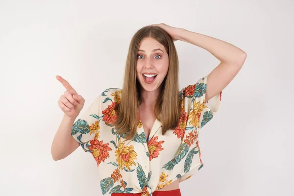 Portrait of young woman with shocked facial expression, showing something amazing on blank space, one hand on her head and pointing with forefinger. OMG concept.