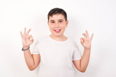 Çekici çocuğun iki eliyle işaret dili göstermesine sevindim. Onaylıyor, neşeli bir ifadesi var, iyimser. Beyaz duvara yaslanarak.