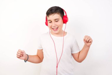 Beyaz duvara karşı günlük kıyafetler giyen, kulaklıkla dans eden ve müzik dinleyen genç bir çocuk..