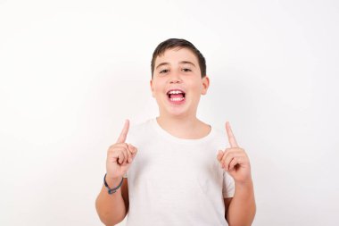 Olumlu ifadeli, kaygısız, her iki işaret parmağıyla işaret eden, günlük kıyafetler giymiş, geniş bir gülümsemesi olan, beyaz arka planda soyutlanmış bir çocuk.. 