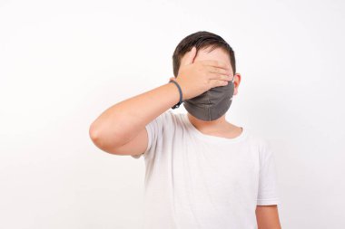 Beyaz arka planda dikilirken, tıbbi maske takan mutlu beyaz çocuk.