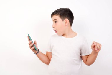 Mutlu bir çocuğun portresi e-posta yoluyla iyi haberler alıyor ve başarıyı kutlarken ayakta duruyor ve beyaz arka planda izole edilmiş cep telefonuna bakıyor..