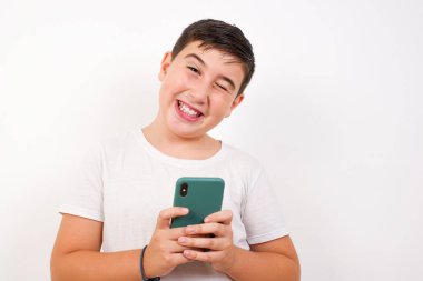 Beyaz arka plan üzerinde duran, günlük kıyafetler giyen, kişisel telefonu kullanan ve kameraya bakıp göz kırpan beyaz çocuk. Flört ve cinsel ilişki kavramı.