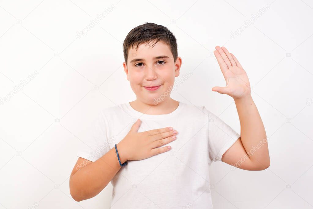 Portrait of happy, sincere young boy raising one arm and hold hand on heart as give oath, pledge, telling truth, want you to believe, standing over white background 