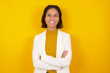 Kollarını kavuşturup gülümseyen esmer kız kameraya bakıp duvara bakıyor. Pozitif kişi