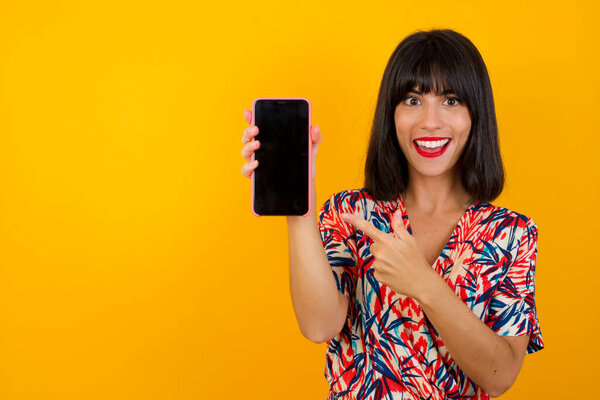 Excited woman pointing with finger at smartphone. Studio shot of shocked girl holding smartphone with blank screen.