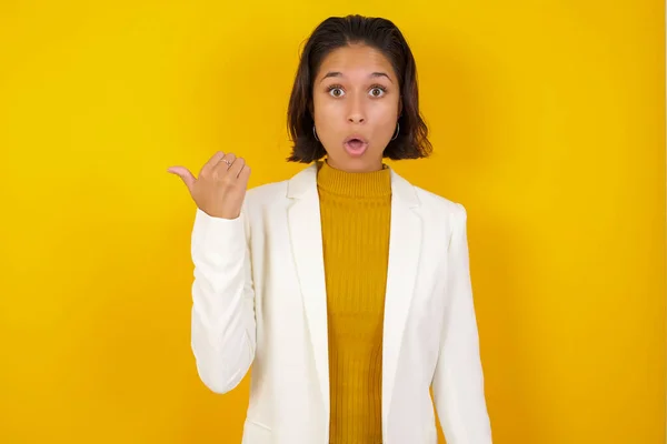 Horizontal shot of pretty young joyful shocked woman  points with thumb away, indicates something on blank wall. Check this out. Ethnicity concept.