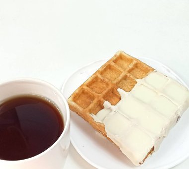 Beyaz krema ya da çikolatayla kısmen kaplanmış Belçika waffle 'ı beyaz bir tabakta oturur. Tabağın yanında koyu bir içecek içeren beyaz bir fincan var, muhtemelen çay ya da kahve. Arkaplan düz beyaz.