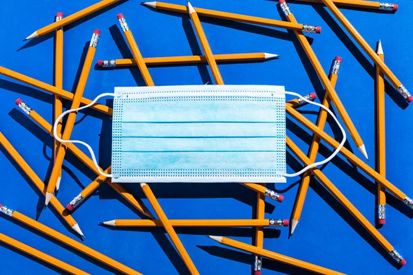 Pencils: pattern of orange pencils and surgical mask from above on blue background. Office, school and protetive concept