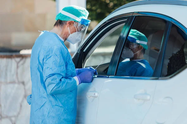 Medical personnel wearing a PPE, performing PCR with a swab in their hand, on a patient inside his car to detect if he is infected with COVID-19