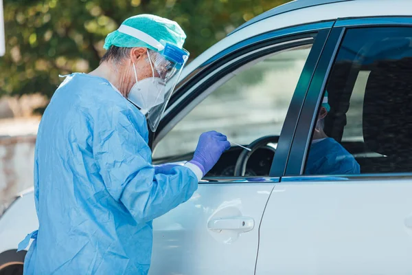 Medical personnel wearing a PPE, performing PCR with a swab in their hand, on a patient inside his car to detect if he is infected with COVID-19