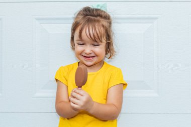 Dondurma yiyen, sarı tişörtlü, beyaz arka plana yaslanmış sevimli sarı saçlı kız.