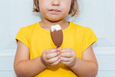 Dondurma yiyen, sarı tişörtlü, beyaz arka plana yaslanmış sevimli sarı saçlı kız.