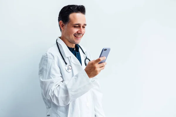 Beyaz önlük ve steteskop giyen siyah saçlı doktor cep telefonuna bakıyor ve beyaz arka planda video görüşmesi yaparken gülümsüyor. Tıp konsepti