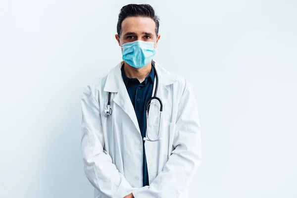 Black haired doctor wearing surgical mask, with white coat and stethoscope looking at camera, on white background. Medicine concept