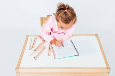 Okuldaki sarışın kız, gülümsüyor, kitaplarla dolu bir masanın yanındaki sandalyeye oturuyor, resim yapıyor, beyaz arka planda. Okul kavramı