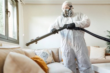 PPE giyen bir adam COVID-19 dezenfektan makinesiyle bir evin oturma odasını dezenfekte ediyor. Salgın sağlık konsepti