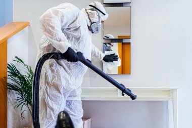 Evde PPE giyen bir adam COVID-19 'dan cam bir masayı dezenfekte ediyor. Salgın sağlık konsepti