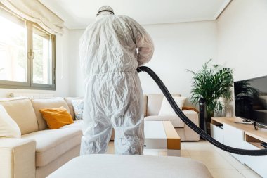 PPE giyen bir adam COVID-19 dezenfektan makinesiyle bir evin oturma odasını dezenfekte ediyor. Salgın sağlık konsepti