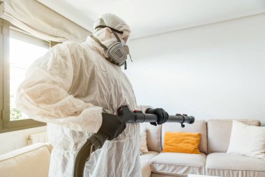PPE giyen bir adam COVID-19 dezenfektan makinesiyle bir evin oturma odasını dezenfekte ediyor. Salgın sağlık konsepti