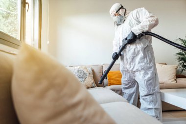 PPE giyen bir adam COVID-19 dezenfektan makinesiyle bir evin oturma odasını dezenfekte ediyor. Salgın sağlık konsepti
