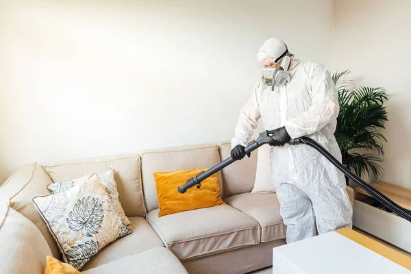 PPE giyen bir adam COVID-19 dezenfektan makinesiyle bir evin oturma odasını dezenfekte ediyor. Salgın sağlık konsepti