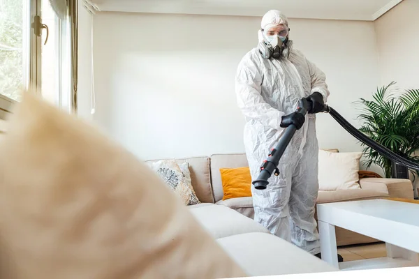 PPE giyen bir adam COVID-19 dezenfektan makinesiyle bir evin oturma odasını dezenfekte ediyor. Salgın sağlık konsepti