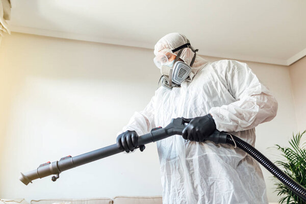 Мужчина в СИЗ дезинфицирует гостиную дома дезинфицирующим аппаратом COVID-19. Пандемия здравоохранения