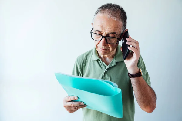 Entrepreneur retired man wearing a green polo shirt and glasses, reading a folder with a report and talking to the mobile