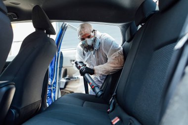 PPE giyen bir adam COVID-19 'dan bir arabanın içini kimyasalla dezenfekte ediyor.