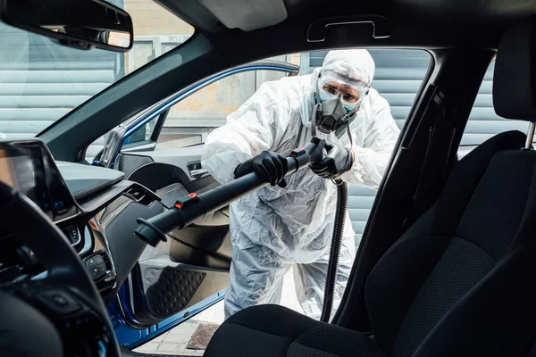 PPE giyen bir adam COVID-19 'dan bir arabanın içini kimyasalla dezenfekte ediyor.